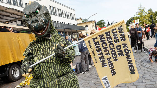 Eine Person im Saurierkostüm hält eine Fahne mit der Aufschrift «Wir können uns die Reichen nicht mehr leisten!». Im Hintergrund Demonstration.