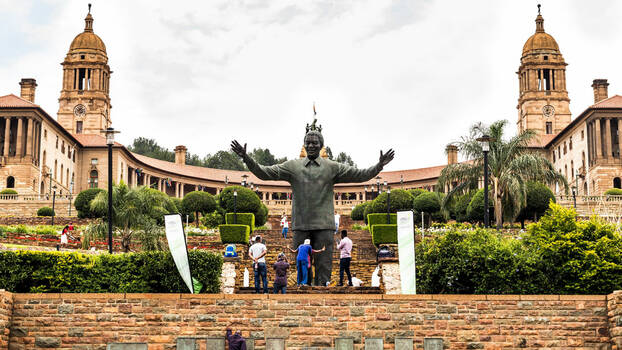 Vor einem großen Sandsteinbau mit, großer Freitreppe, zwei Türmen und Säulengang steht eine große Statur von Melson Mandela mit ausgebreiteten Armen. Davor stehen Menschen und fotografieren